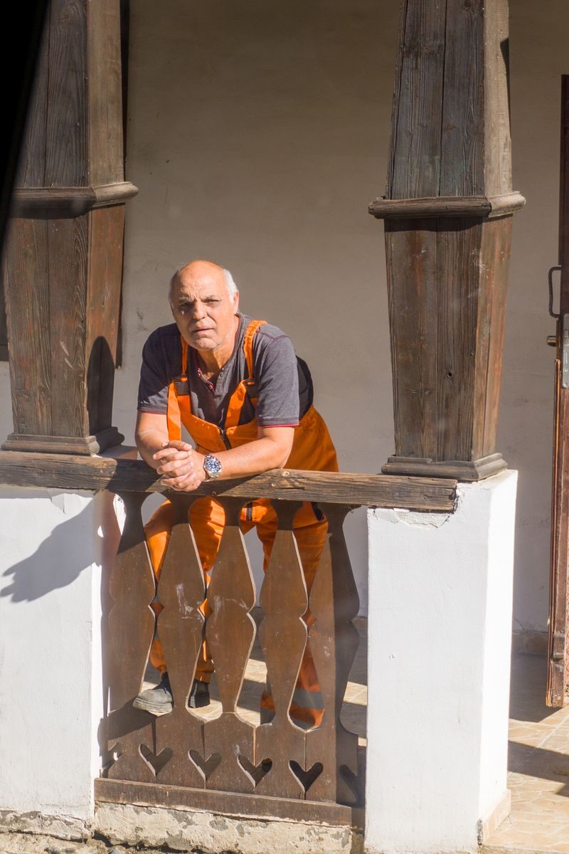 An elderly man in work overalls leaning on a vintage wooden railing.