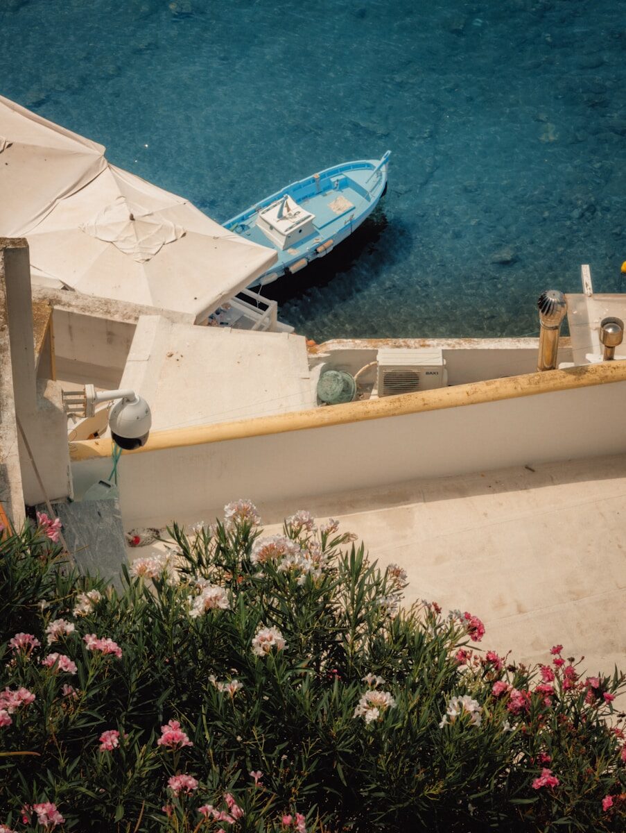 A blue boat moored in clear water beside a white terrace with flowers.
