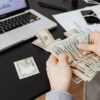 Person counting dollar bills at desk with laptop and financial charts, symbolizing finance management.