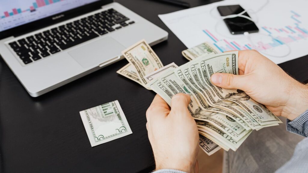 Person counting dollar bills at desk with laptop and financial charts, symbolizing finance management.