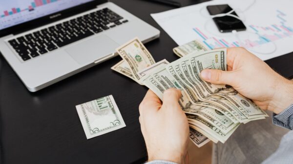 Person counting dollar bills at desk with laptop and financial charts, symbolizing finance management.