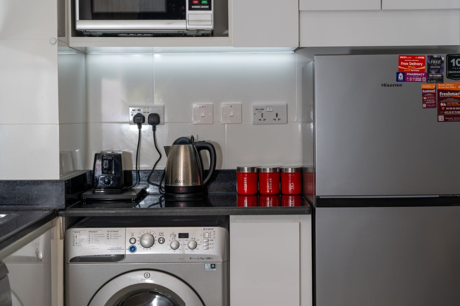 Stylish modern kitchen with stainless steel appliances and red accents, highlighting a washing machine, fridge, and countertop electronics.