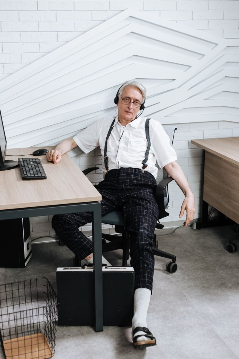 Senior man in a white shirt and headphones relaxing in an office chair by a desk.