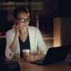 Woman in glasses working late at night on laptop, holding coffee, making phone call.