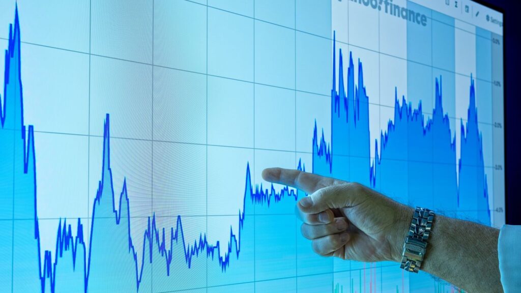 A hand points to a financial graph on a large screen, indicating market trends.