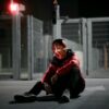 A person wearing an Iron Man helmet sits on the ground at night in an urban setting.