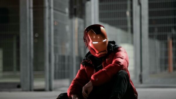 A person wearing an Iron Man helmet sits on the ground at night in an urban setting.