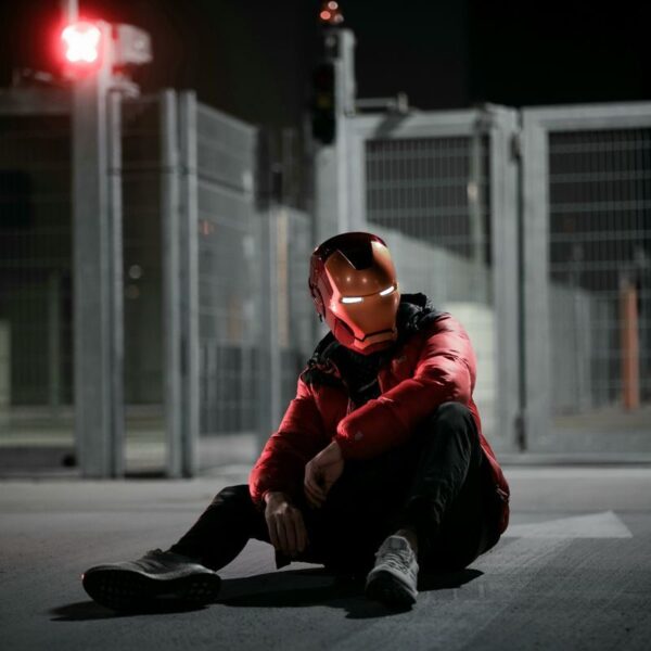 A person wearing an Iron Man helmet sits on the ground at night in an urban setting.