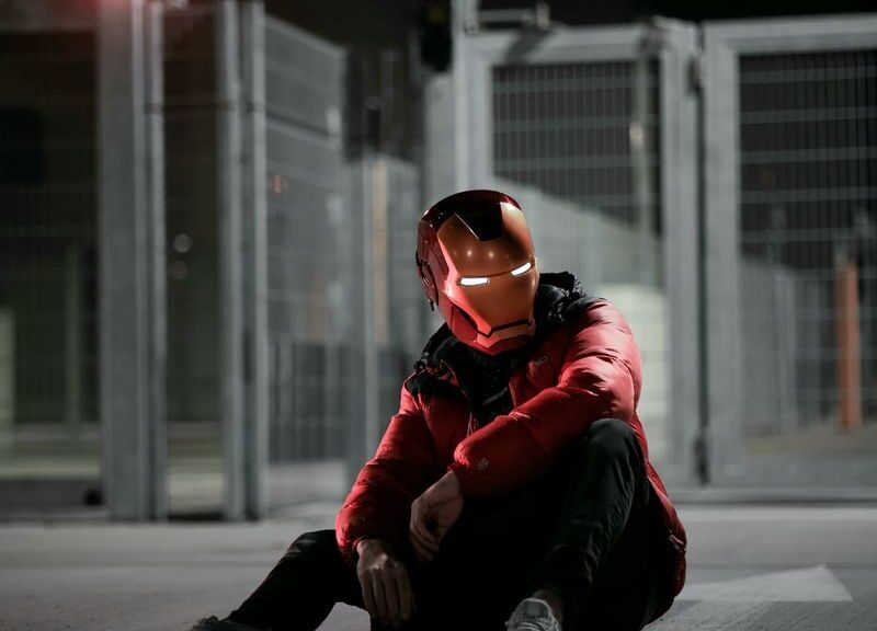 A person wearing an Iron Man helmet sits on the ground at night in an urban setting.