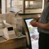 Unrecognizable male in casual clothes standing with money near cash register in grocery store while making purchase near glass showcase