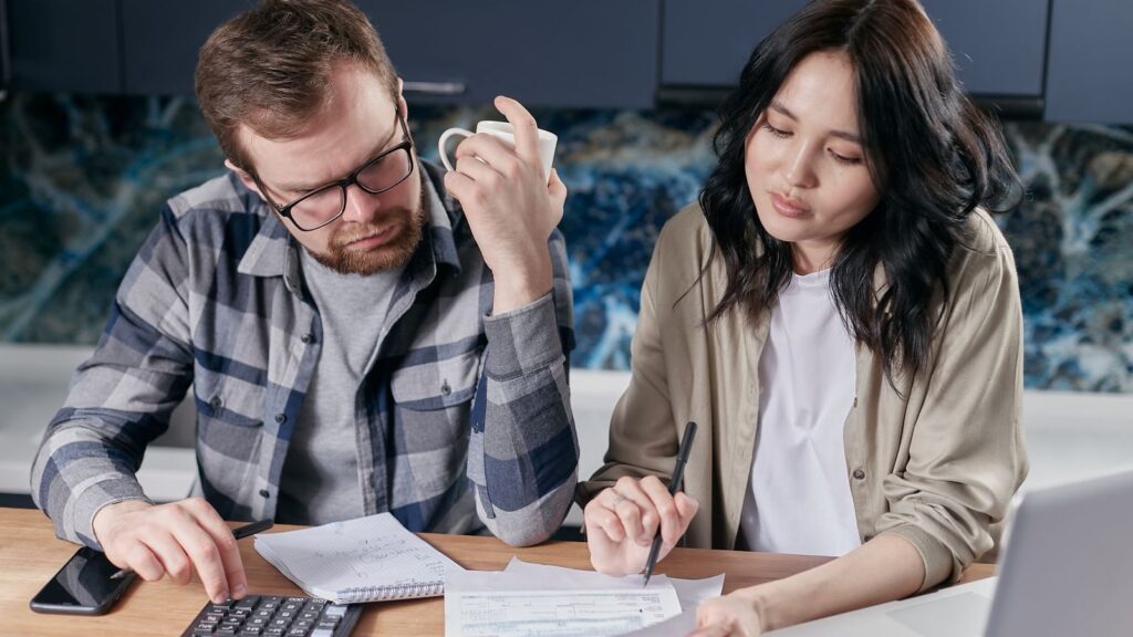 A couple reviewing household bills and budget using a calculator and laptop at their kitchen table.