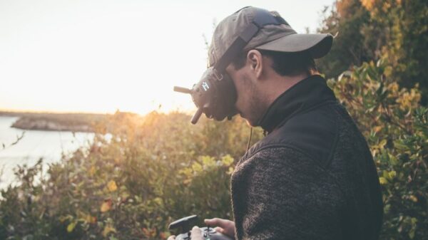 Adult man using VR goggles and a drone controller in a scenic outdoor setting during sunset.