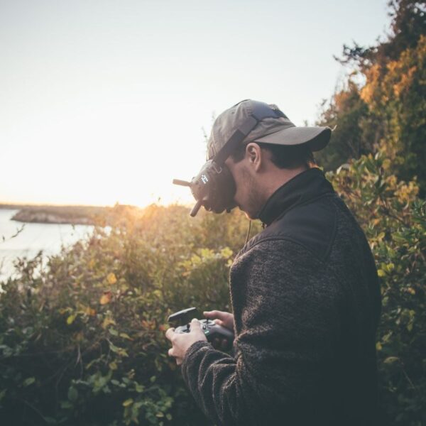 Adult man using VR goggles and a drone controller in a scenic outdoor setting during sunset.