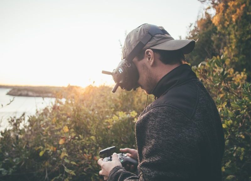 Adult man using VR goggles and a drone controller in a scenic outdoor setting during sunset.