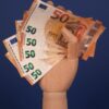 Wooden hand holding multiple euro banknotes against a blue backdrop, symbolizing finance and wealth.