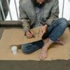 Person sitting on cardboard barefoot, writing a sign with a paper cup nearby.