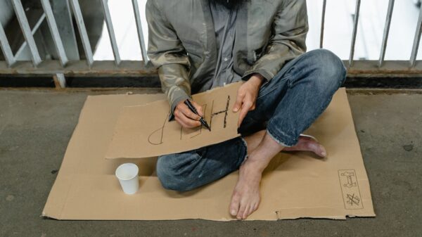 Person sitting on cardboard barefoot, writing a sign with a paper cup nearby.