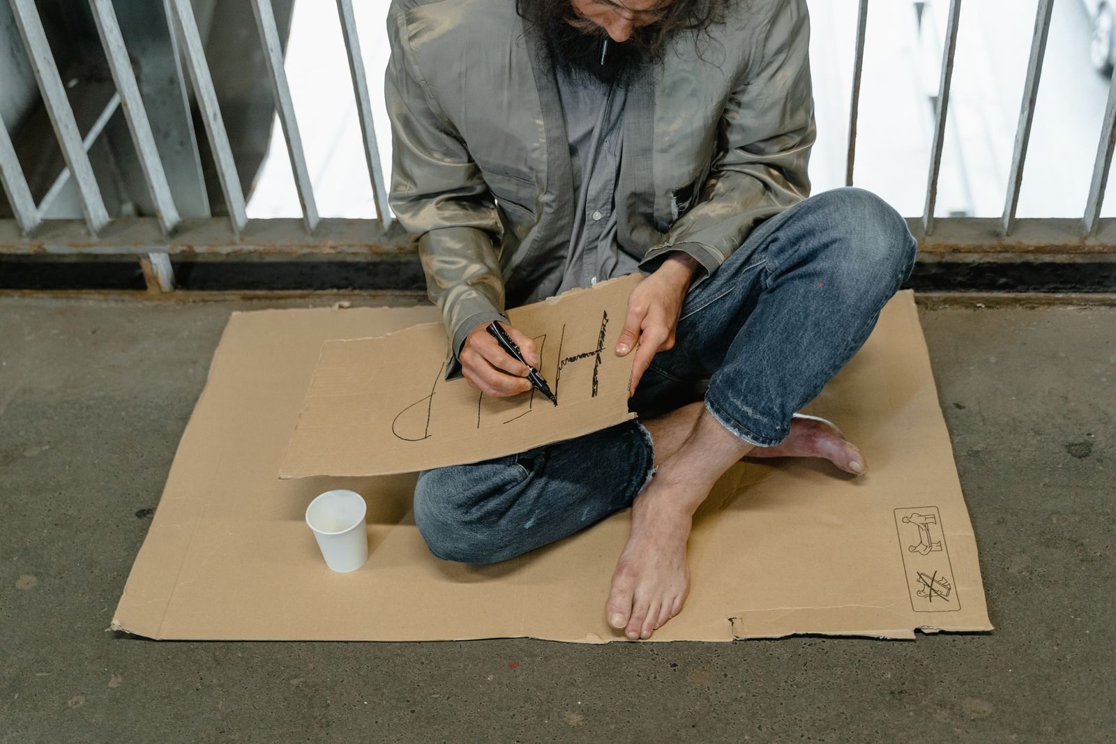 Person sitting on cardboard barefoot, writing a sign with a paper cup nearby.