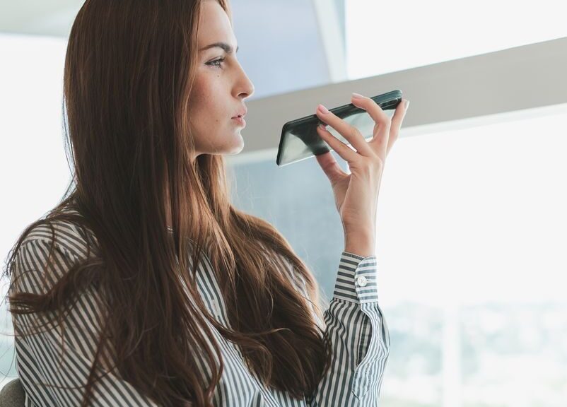 Young businesswoman using voice assistant on her phone in a modern office setting.