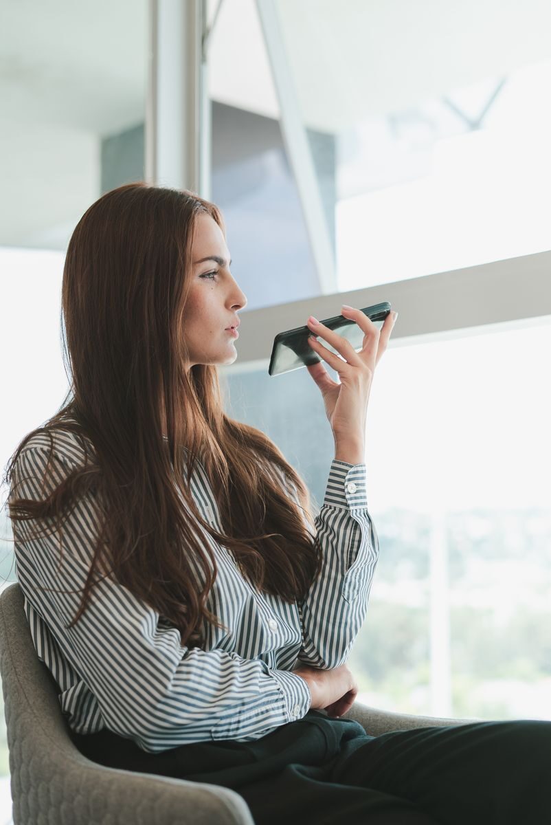 Young businesswoman using voice assistant on her phone in a modern office setting.