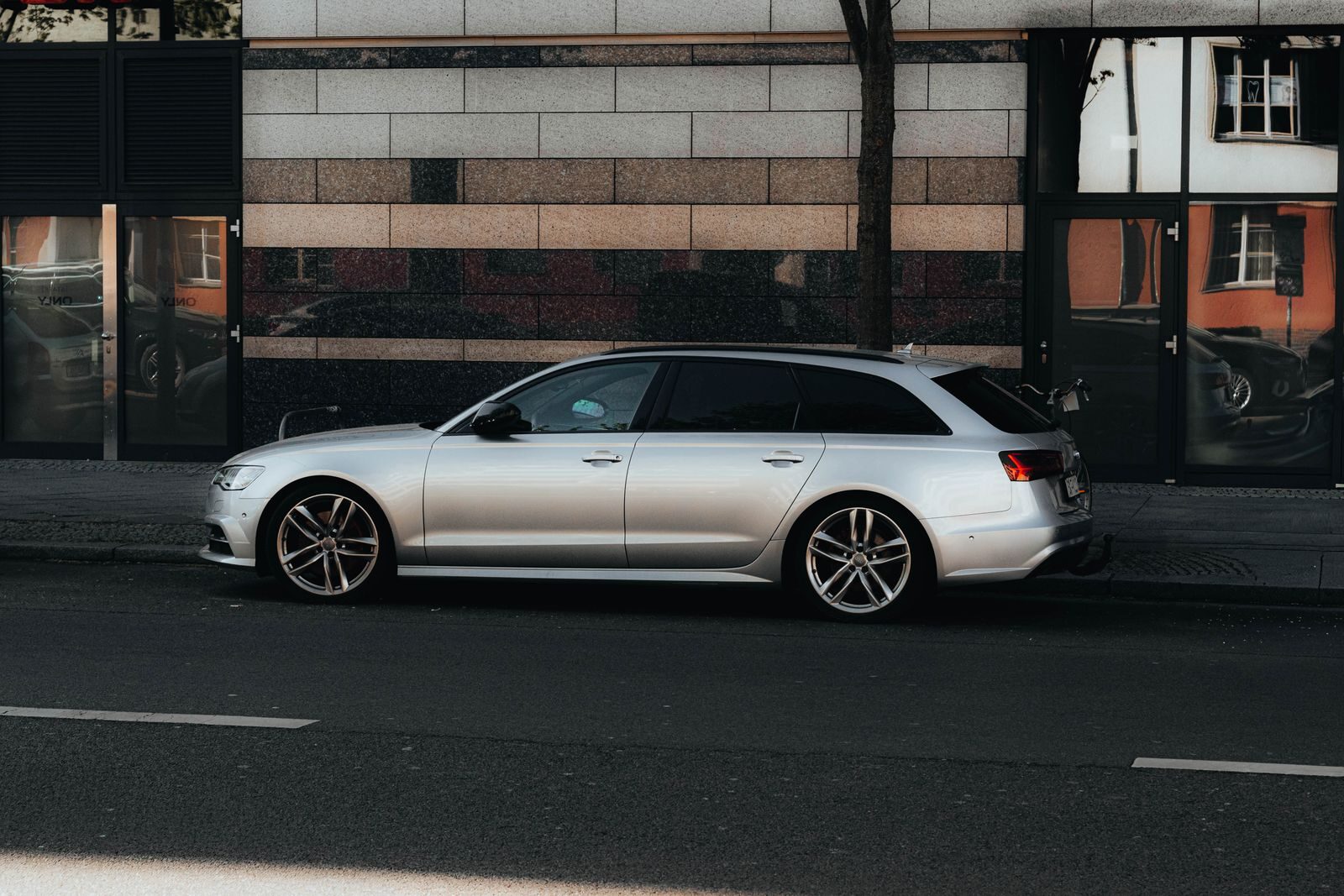 A silver luxury car parked on a Berlin street with modern architecture reflecting in city windows.
