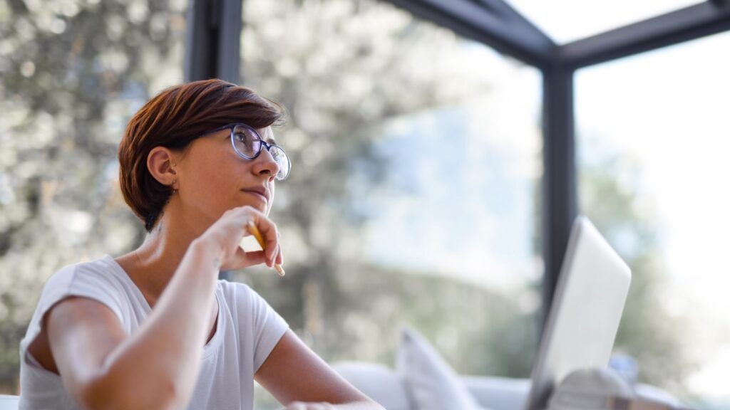 Thoughtful woman with glasses and short hair looking out window in a modern room.