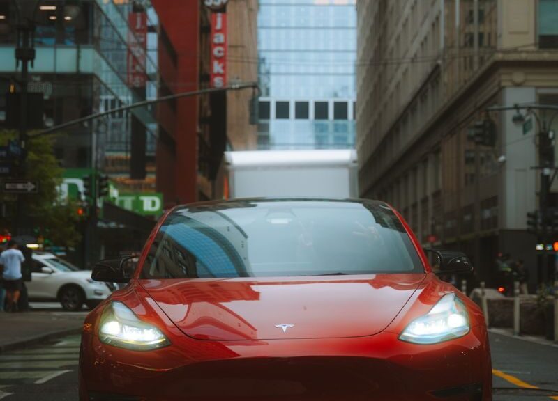 Red Tesla Model 3 car on an urban street with skyscrapers and cityscape view.