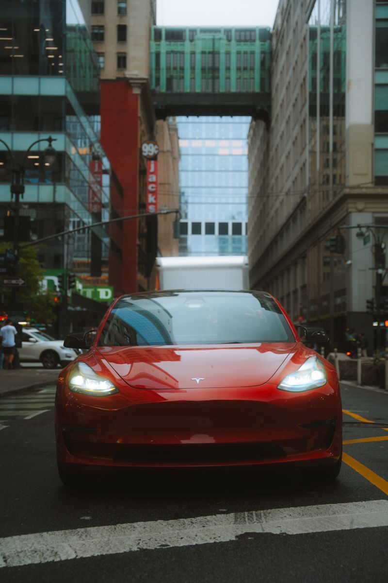Red Tesla Model 3 car on an urban street with skyscrapers and cityscape view.