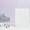 Minimalist image of a white shopping bag and mini cart on a soft pastel background.