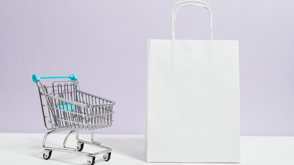 Minimalist image of a white shopping bag and mini cart on a soft pastel background.