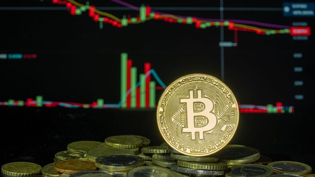 Bitcoin coin among stacked coins against a backdrop of a fluctuating financial market chart.