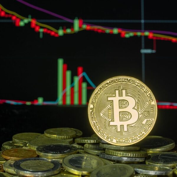 Bitcoin coin among stacked coins against a backdrop of a fluctuating financial market chart.