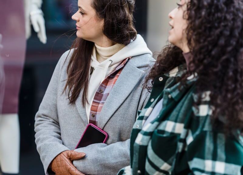 Pensive women friends in casual clothes standing with shopping bags near shop on street in daytime