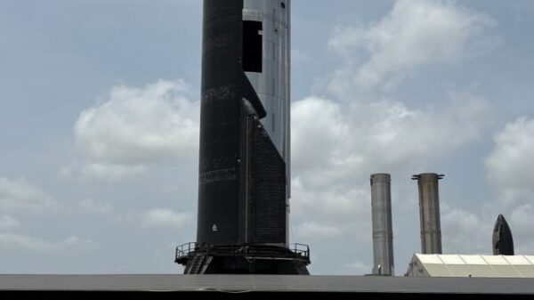 Tall rocket at launch pad with parked cars below under a cloudy blue sky