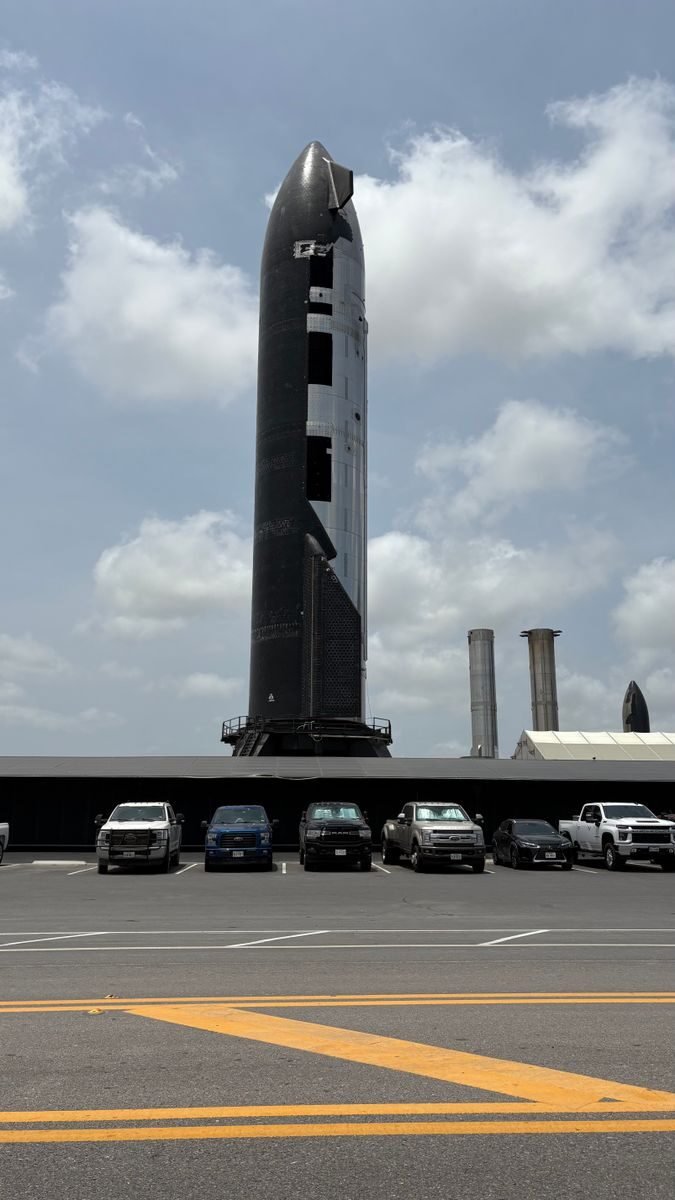 Tall rocket at launch pad with parked cars below under a cloudy blue sky