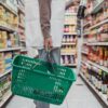 Side view of a woman shopping in a supermarket aisle holding a green basket and a drink bottle.